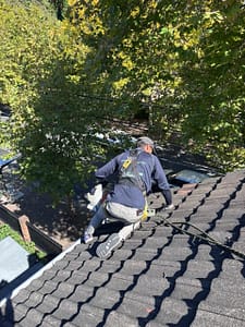 Professional roof technician wearing a full safety harness while using a leaf blower on a steep tiled roof during a gutter cleaning Sydney service, safely removing leaves and roof debris