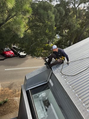 Gutter Cleaning Sydney Professional technician carrying out gutter cleaning Sydney services, removing leaves and debris from a steep Colorbond roof while using certified height safety equipment.